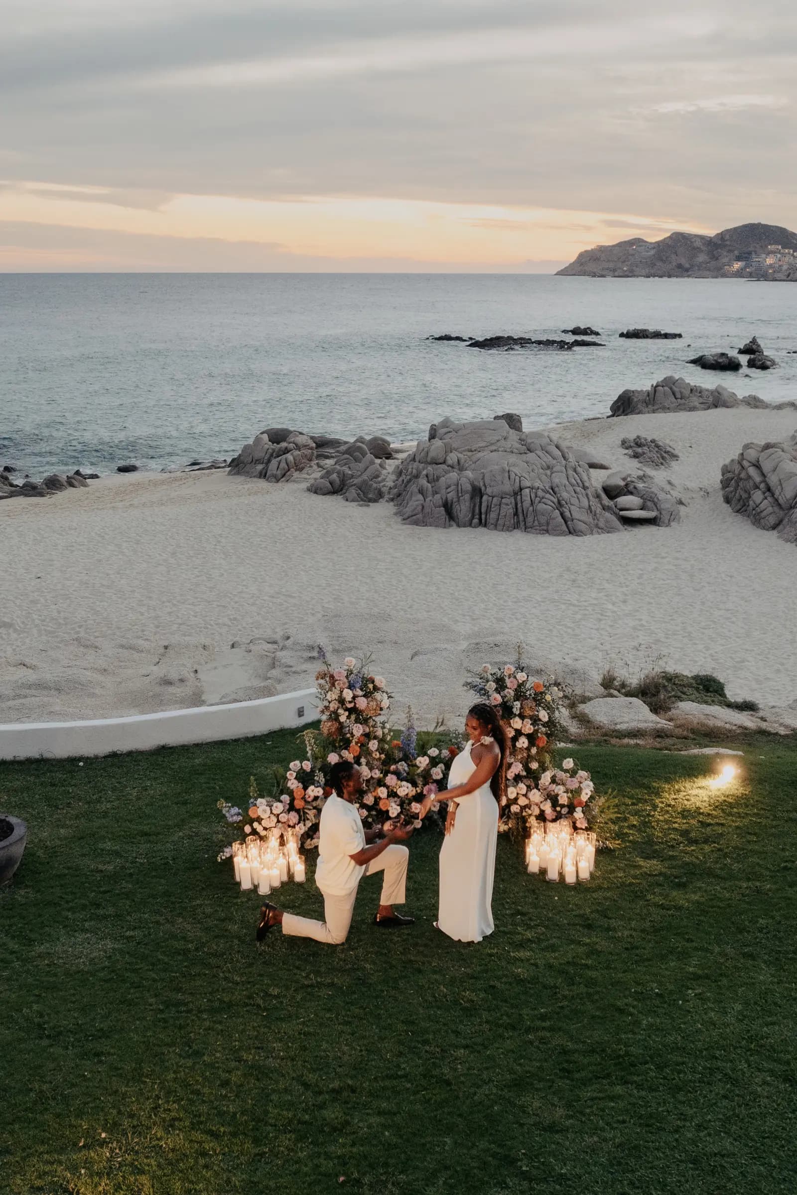 Olugbenga on one knee proposing to Temiloluwa at sunset by the sea in Cabo, black and white
